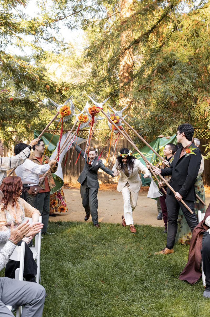 queer couple walk down the aisle together through breakaway banner, under a halberd arch