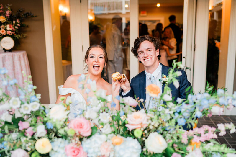 A smiling bride and groom sit behind a table decorated with colorful flowers, holding and displaying desserts together at their wedding reception. A group of people is visible in the background.