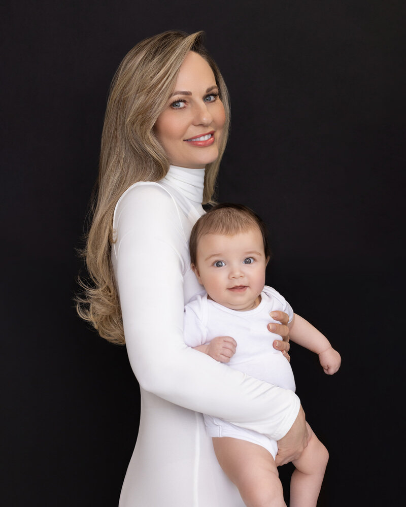 London Studio portrait of mother holding baby and both smiling to the camera dressed in white outfits on the  black background.