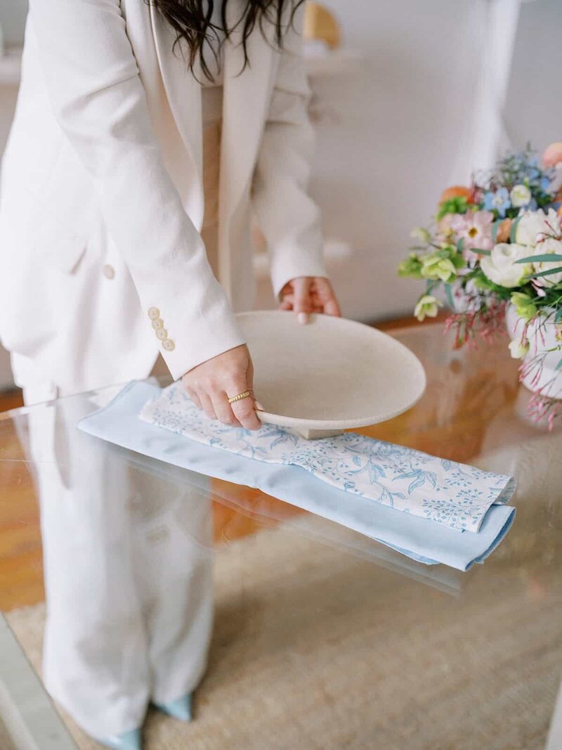 A wedding planner client of a Showit website designer in a white suit sets a table with a white plate and a folded blue embroidered napkin on a glass surface. A bouquet of colorful flowers adorns the table.