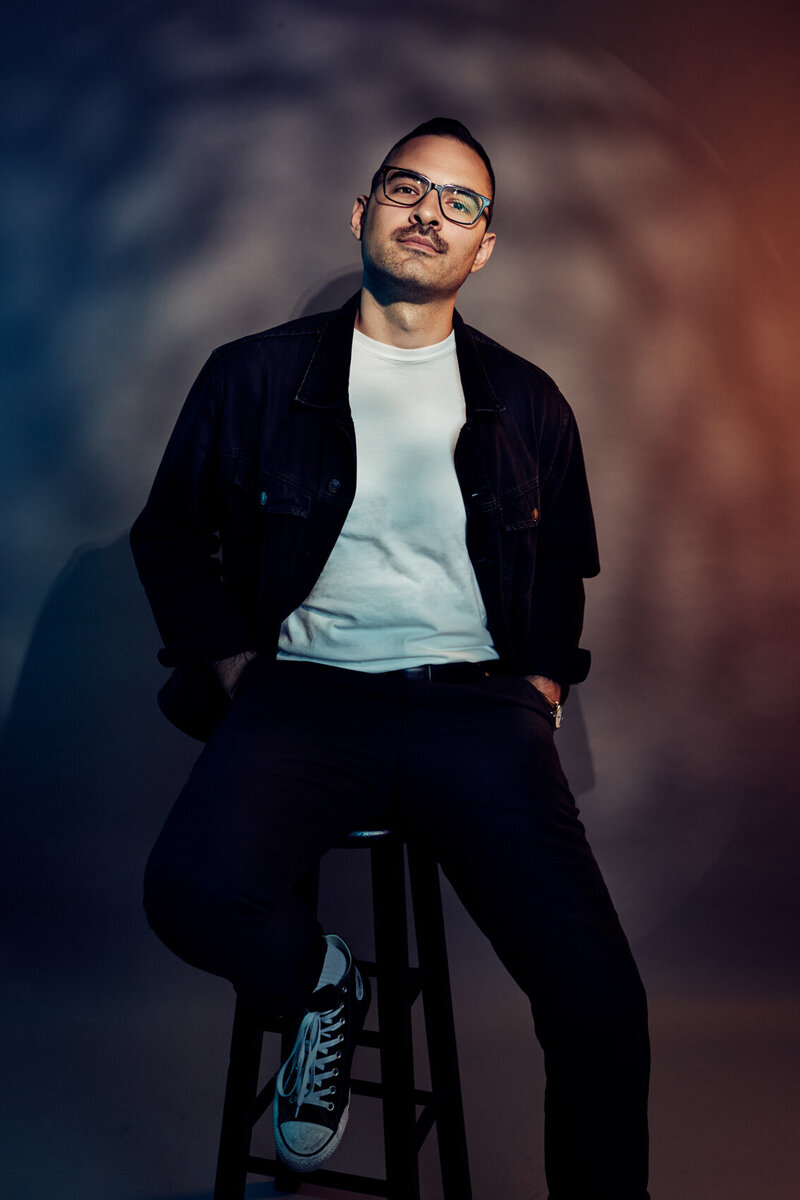 Studio portrait of Mike Moretti seated on a stool, wearing a dark jacket and light shirt, with dramatic gradient lighting and a confident pose, captured by Ylva Erevall.