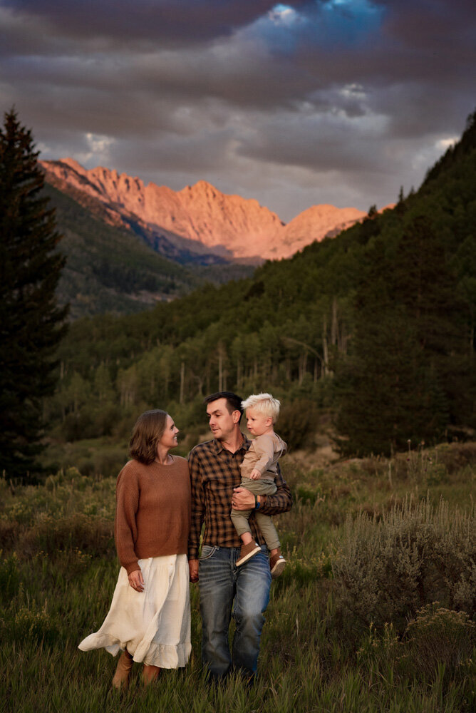 In a mountain meadow a mom and dad  walk together while holding their son