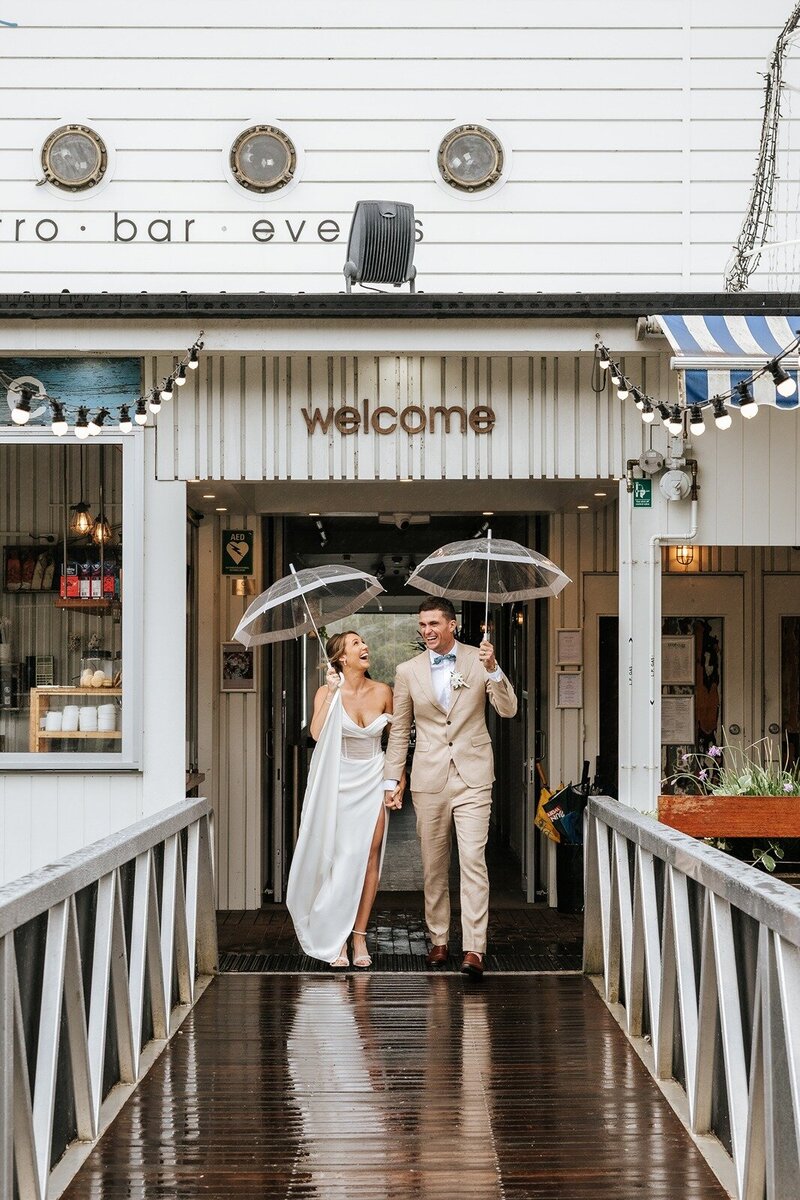A bride and groom walking in the rain with umbrellas on the deck at Noosa Boathouse