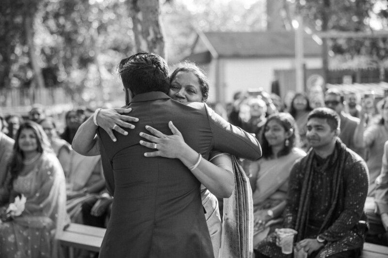 A mom embraces her son warmly in a ceremony setting, surrounded by guests in elegant attire, radiating joy and celebration.