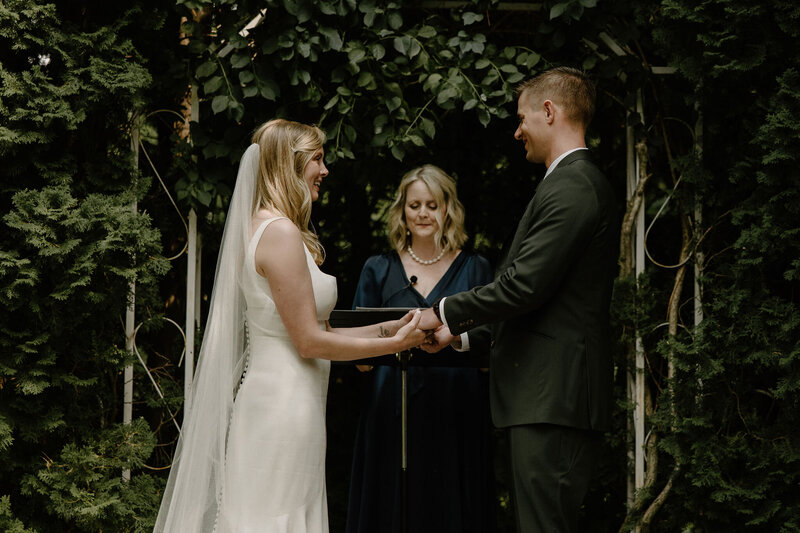 Dark and vintage inspired wedding photography session by Morgan Ashley Lynn Photography of the ceremony where the bride and groom are holding hands saying their vows, smiling in front of the officiant, in front of a forest