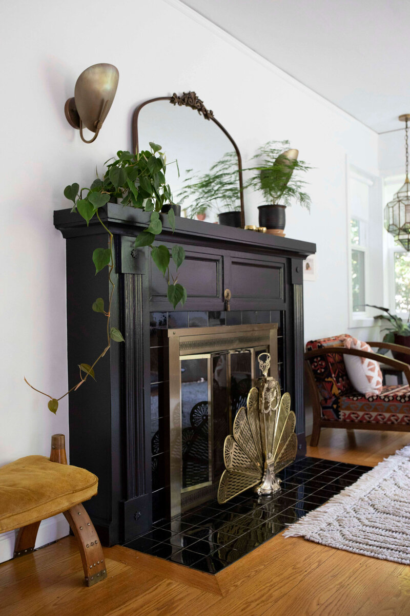 A living room featuring an organic modern aesthetic with a black-painted fireplace. The mantel is decorated with potted plants a vintage mirror, and a gold peacock-style fireplace screen adds a touch of glamour. 