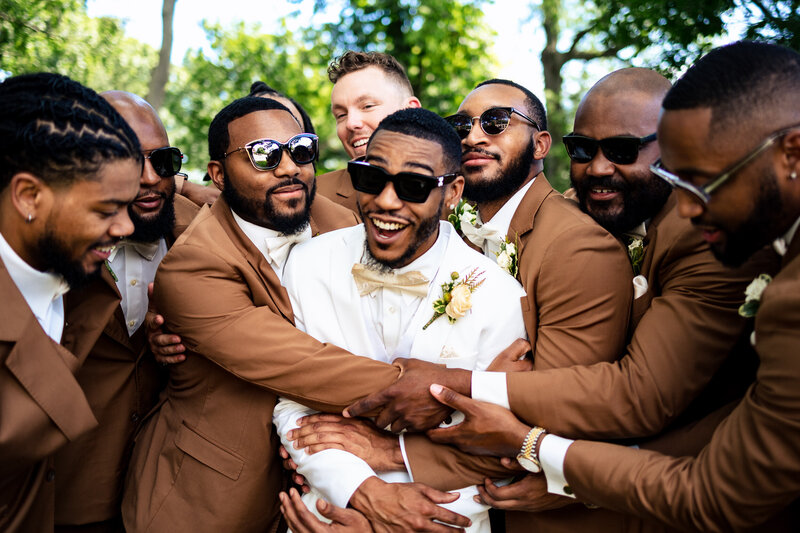 Groomsmen surrounding the groom giving him a hug as he laughs and smiles outside of the Toledo Museum of Art