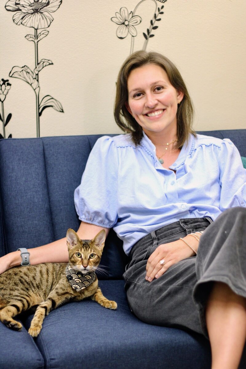 Two people sit on an orange couch, their arms resting near a stack of books and magazines. Both wear light-colored clothing. This calm scene could be from a session with an eating disorder therapist in Orange County. Only their lower faces and bodies are visible.