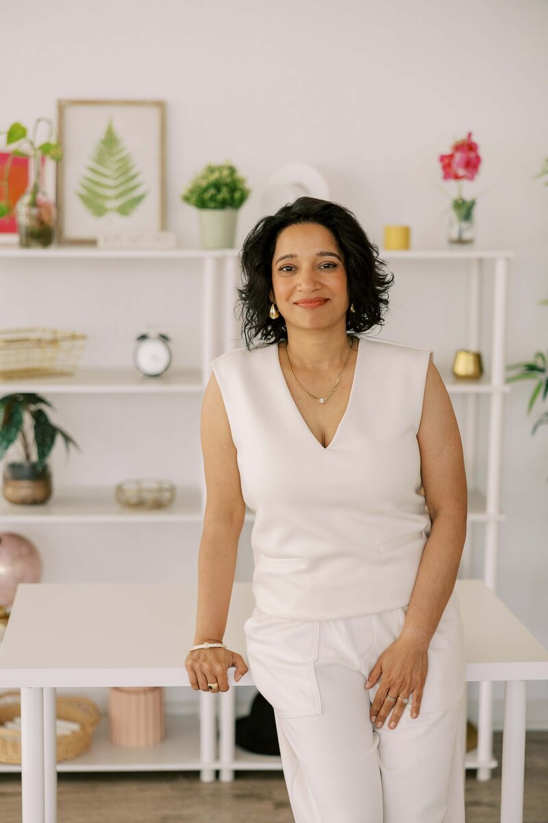 Woman leaning casually on a table, wearing an all white outfit and smiling at the camera with a soft smile