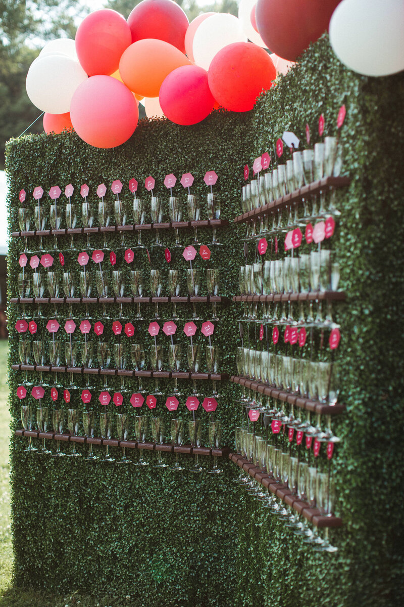 A large boxwood wall with rows of filled champagne flutes that have  fuchsia escort cards in them sits on a lawn with a  coral balloon garland on top, outside at Rockport Barn in Thousand Islands, designed by Frid Events.