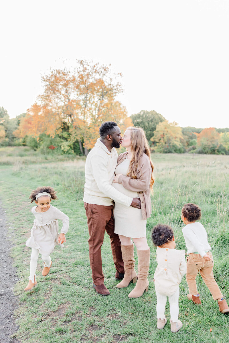 Mom and dad kiss in a meadow while their three children run around them