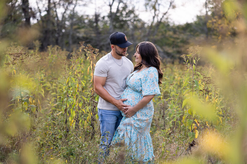 Expecting couple looking at one another with hands on mom's belly while they stand in a field at sunset. 