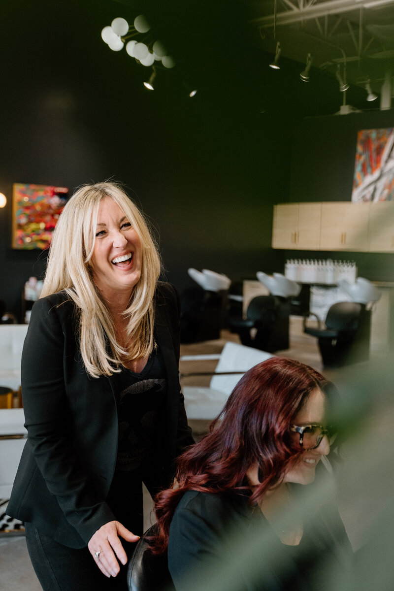 Brand photography portrait of hair stylist and colorist smiling and laughing with client in salon chair.