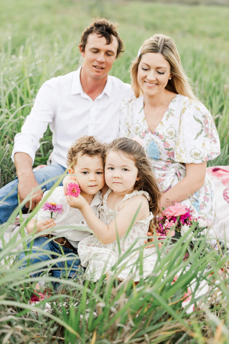 A little girl cuddles her little brother a they sit in front of their parents.  The little girl has on a beautidul floral dress while the little boy sis dressed in a pair of jeans and white shirt.