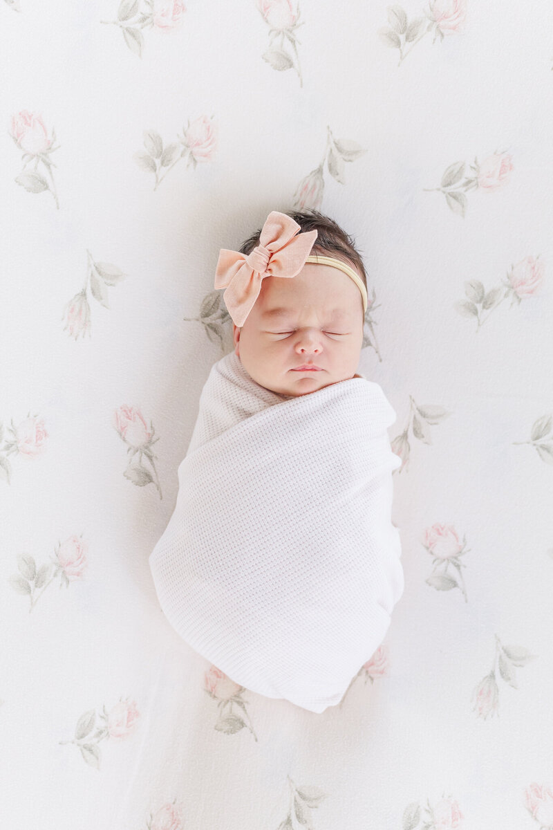 newborn baby girl wrapped in pink swaddle sleeping quietly with pink bow laying on light pink rose colored background