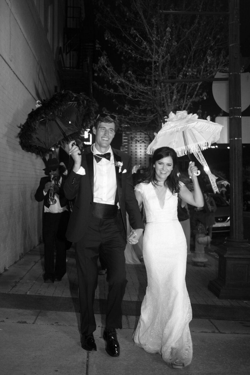 A bride and groom, captured by wedding photographer, stroll down the street hand-in-hand, sheltered under whimsical umbrellas.