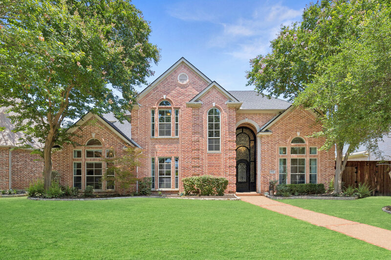 Stunning brick exterior at 4947 Sandestin Drive with impressive curb appeal, featuring mature shade trees, manicured landscaping, and a grand arched iron front door flanked by tall, elegant windows that set the tone for this beautifully maintained two-story home.