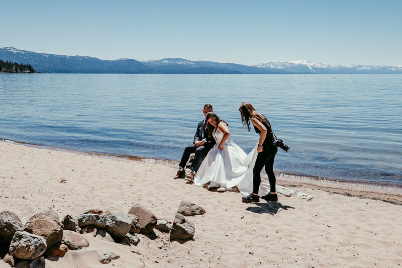 Lake Tahoe elopement photographer Kristin Smith helping a bride with her dress during beach wedding photos by the lake with snow-capped mountains in the background.