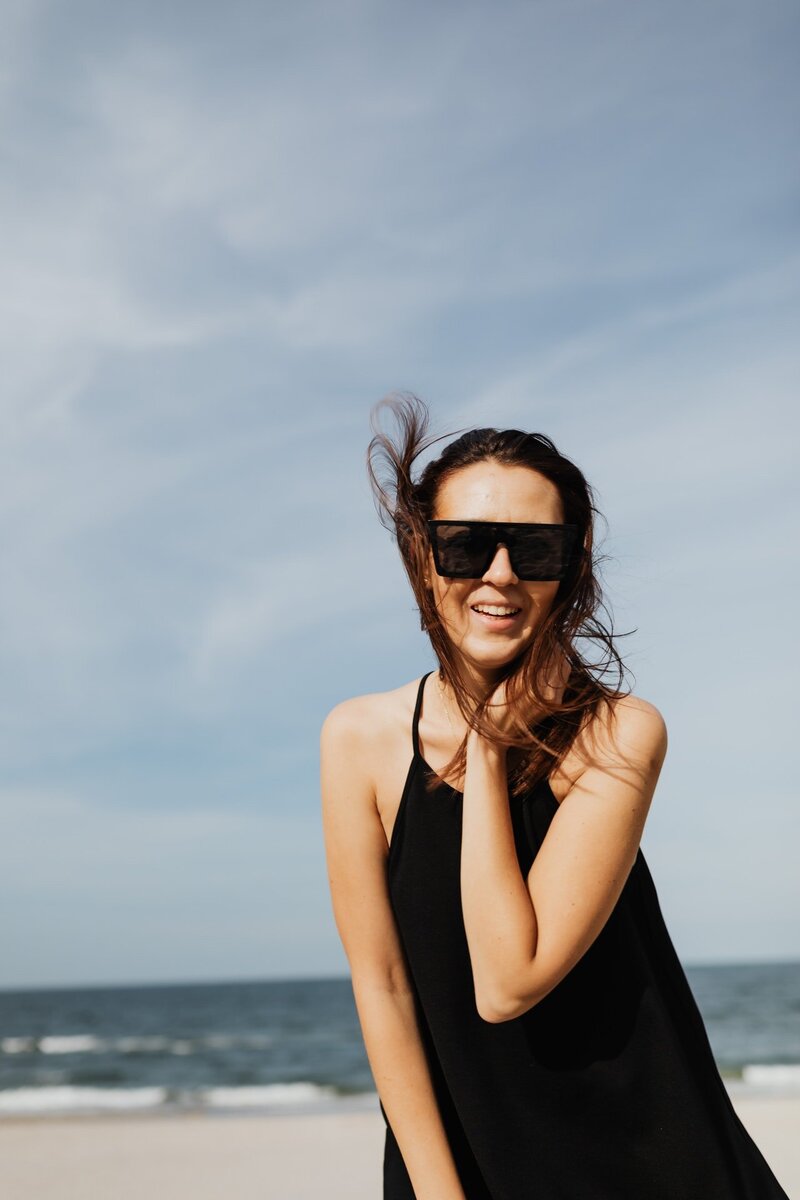 A photo of a woman outside at the beach wearing black sunglasses and a black dress smiling.