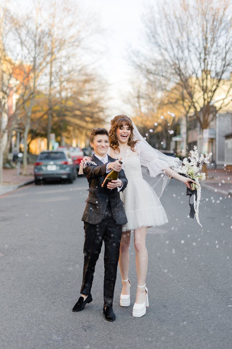 Nonbinary newlyweds celebrating in the street in Georgetown DC popping a spraying champagne bottle while laughing and smiling