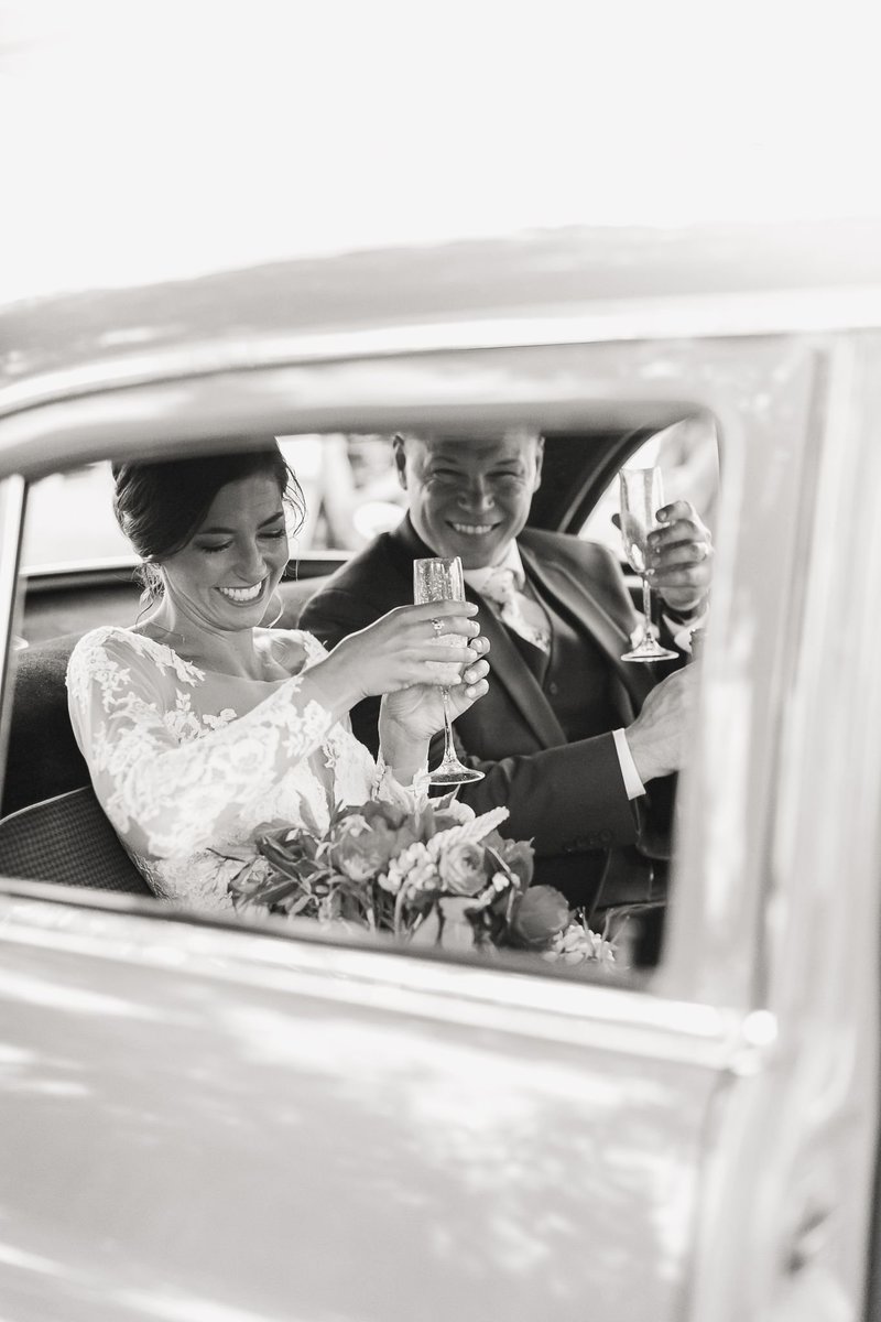 A black and white portrait of a bride and groom toasting champagne in the back seat of a vintage car.