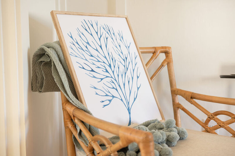 a framed canvas of a blue marine plant watercolor leaning on a blanket draped over a boho rattan chair