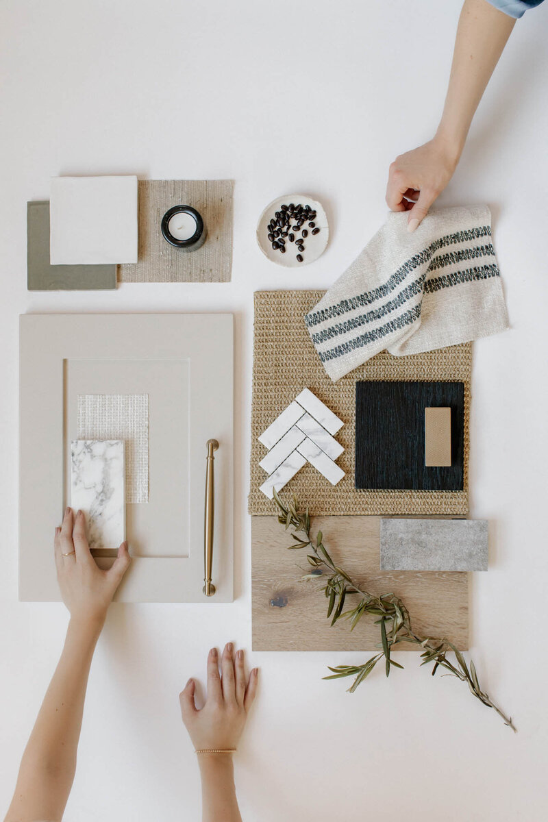 Flatlay photo of design materials for inspiration, the theme is soft textures, beige, with dark accents. Some of the materials are small white marble tiles, bronze handles, and woven fabrics