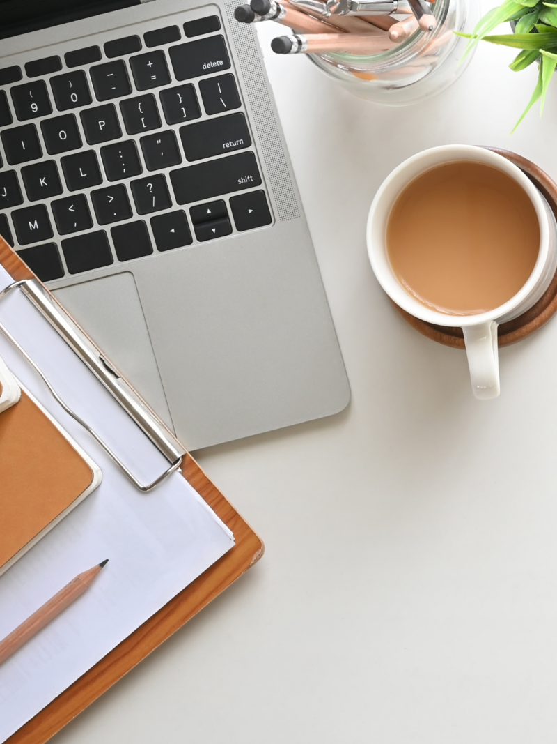 Flat lay of a laptop, clipboard with notes, coffee cup, and jar of pencils on a white desk.