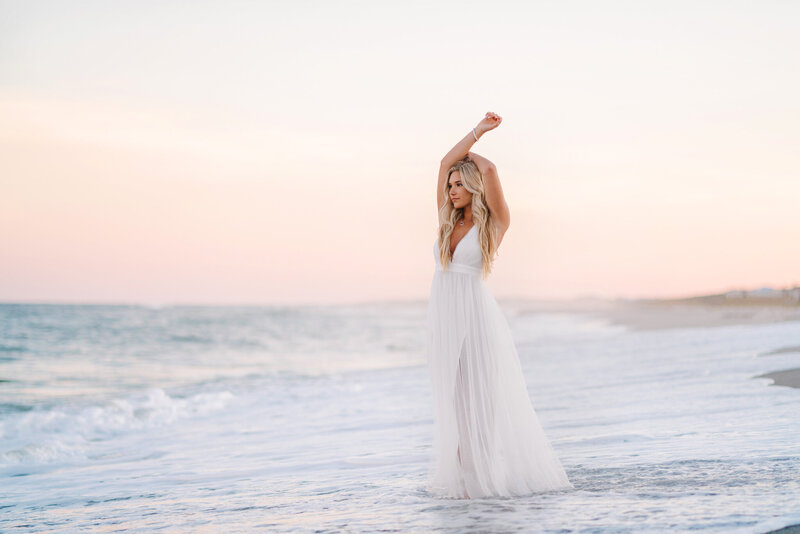 High school senior girl in white dress posing with arms up during sunset on the beach in Charleston, SC
