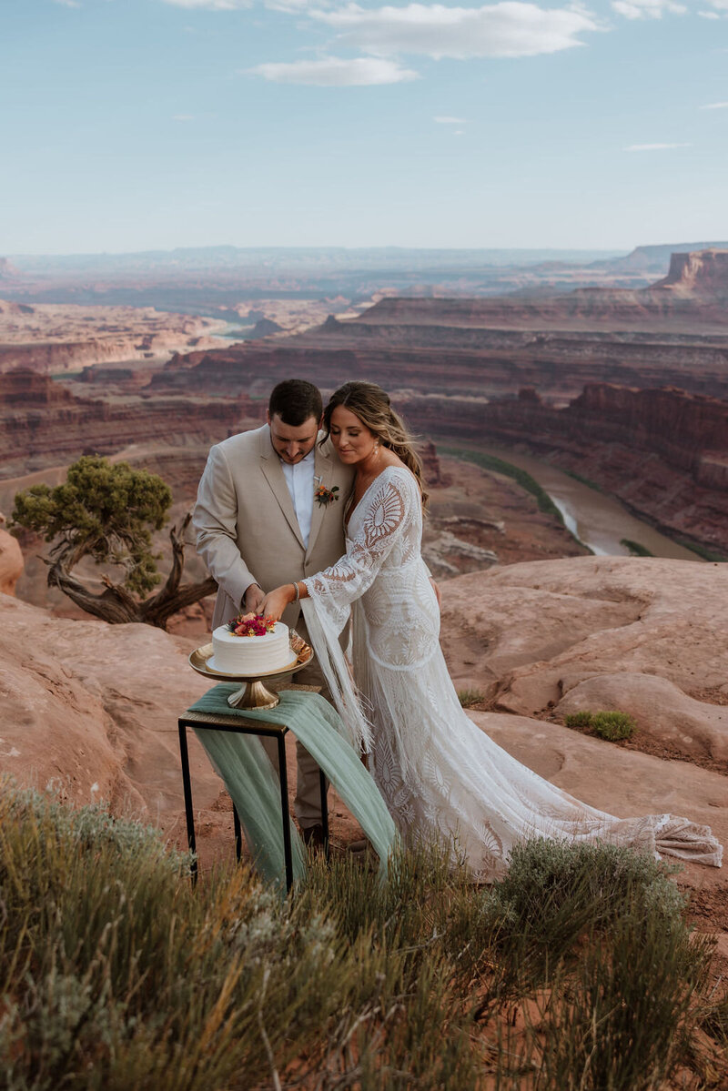 bride and groom cutting their cake on a small table at the edge of a cliff in the desert