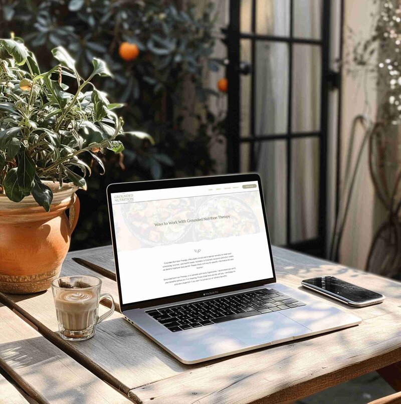 A laptop sits on an outdoor table surrounded by plants, with a steaming cup of coffee nearby displaying a website for a nutritionist in Colorado.