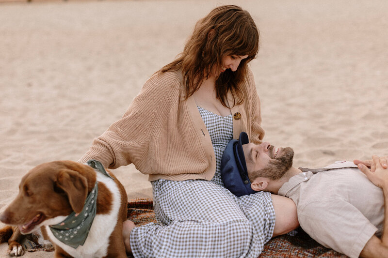 A woman sits on a sandy beach with a dog beside her, enjoying a relaxed moment with a companion.