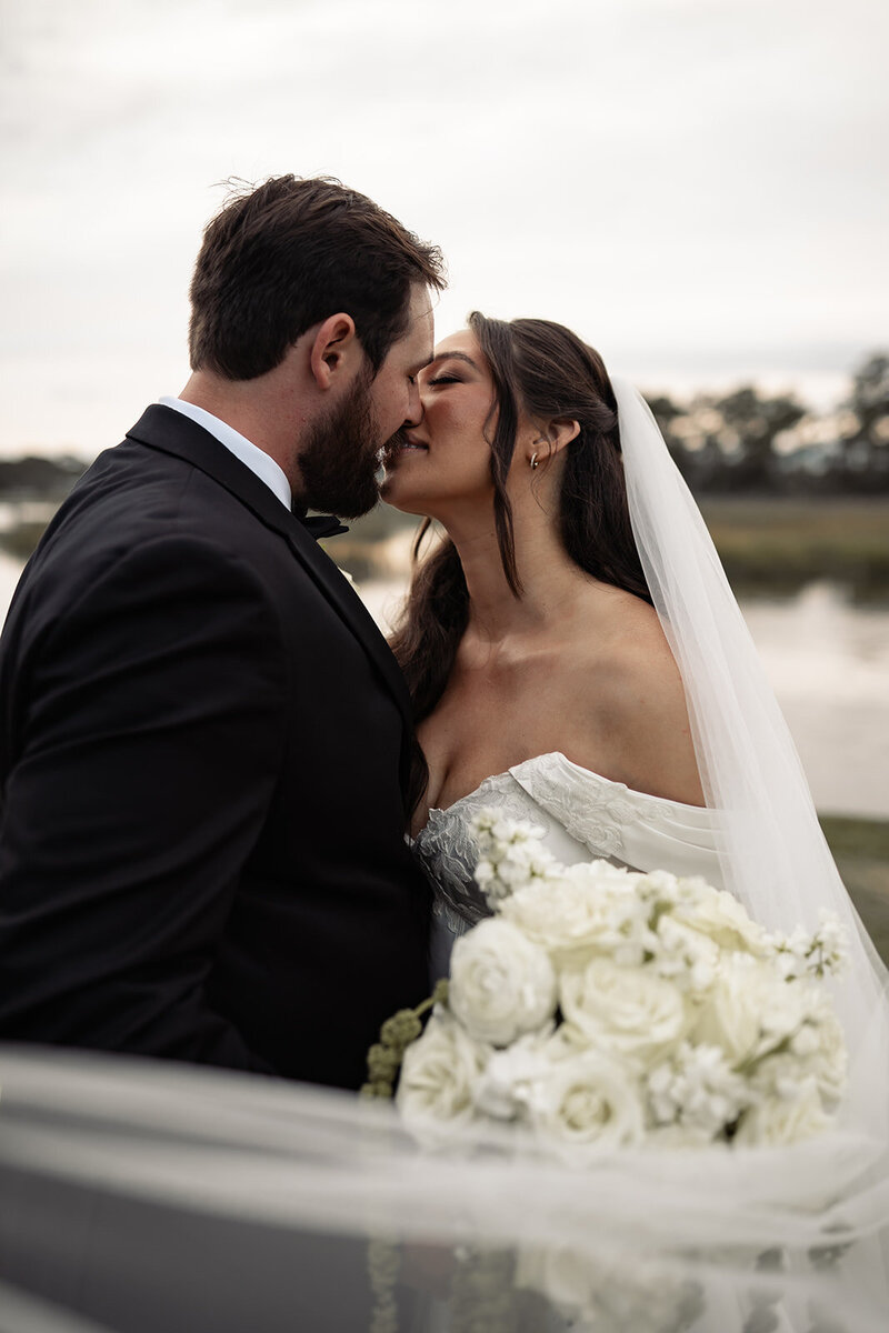 Bride and groom real close for a kiss with their lips almost touching. bride is holding her bouquet in between them with her veil flowing around the bouquet and into the camera frame