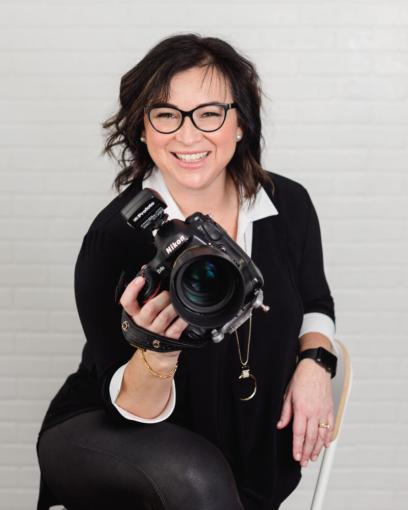 a branding photo of Ottawa Headshot Photographer Elenora Luberto of JEMMAN Photography COMMERCIAL wearing black, sitting in a chair holding her camera