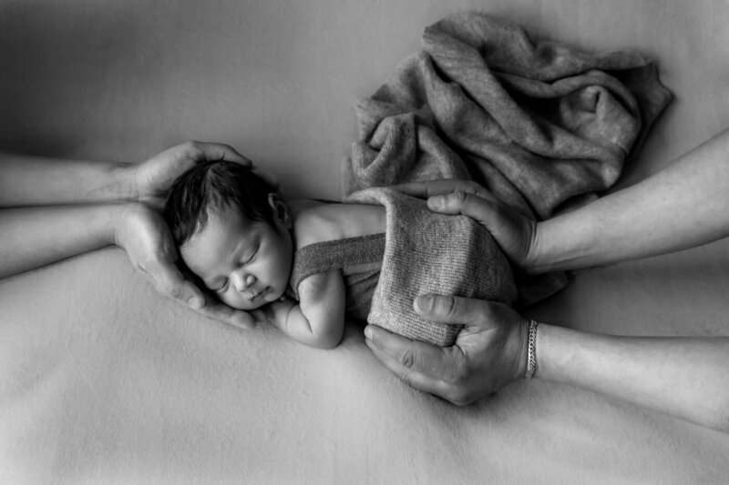 Black and white photo of a newborn baby sleeping on a soft blanket, gently cradled by two pairs of adult hands