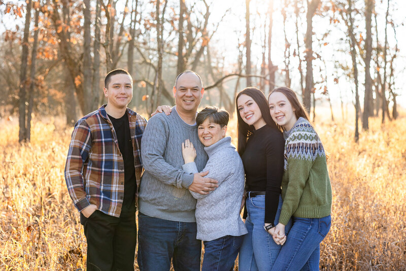 Family portrait of mother, father, and three adult children smiling while standing among the trees. 
