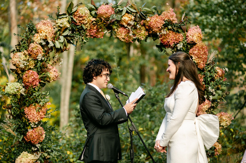 Groom sharing his vows to his bride with flower wedding arch in background