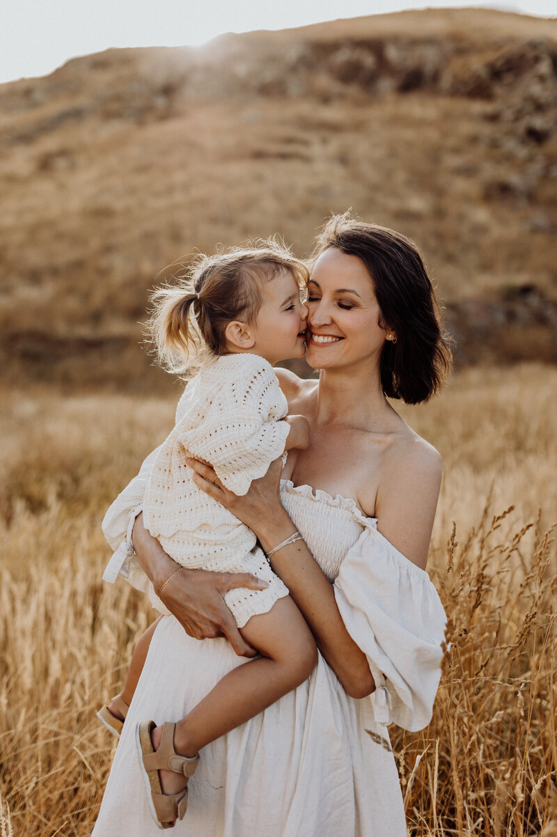 A mother holds her baby wearing a dress from lucy janes client wardrobe in the christchurch hills in a field of long grasses
