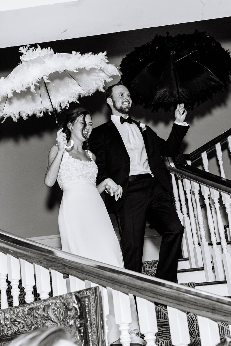 Black and white photo of  bride and groom walking down staircase holding feather umbrellas