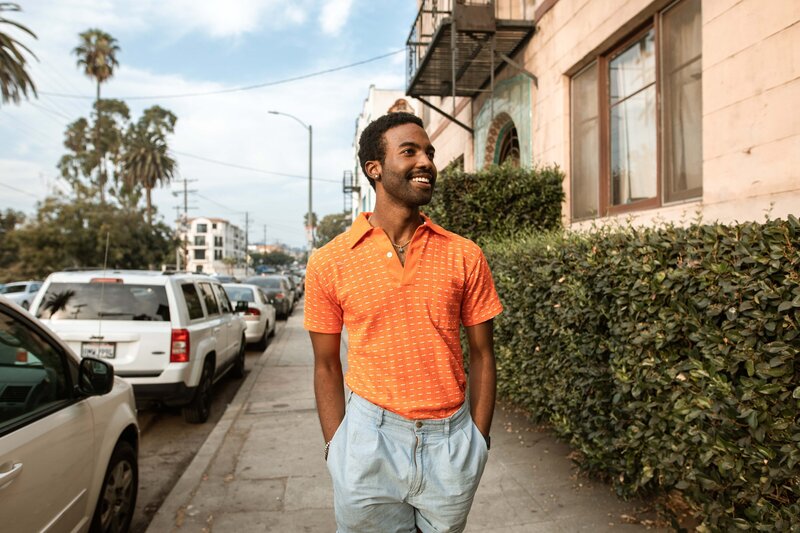 This image shows a young, masculine-presenting person of color walking down a sidewalk toward the camera. The sidewalk is lined by parked cars on one side and green bushes on the other.