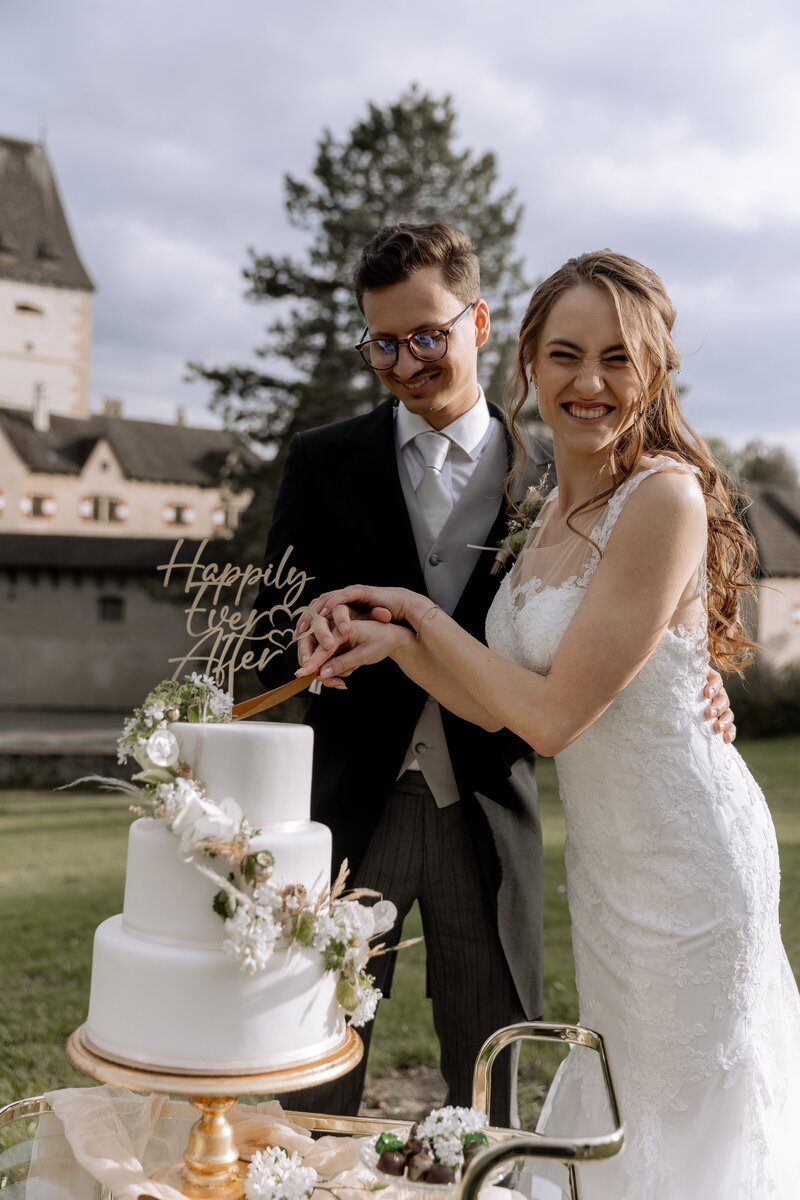 Braut und Braeutigam schneiden weisse Hochzeitstorte an und lachen dabei