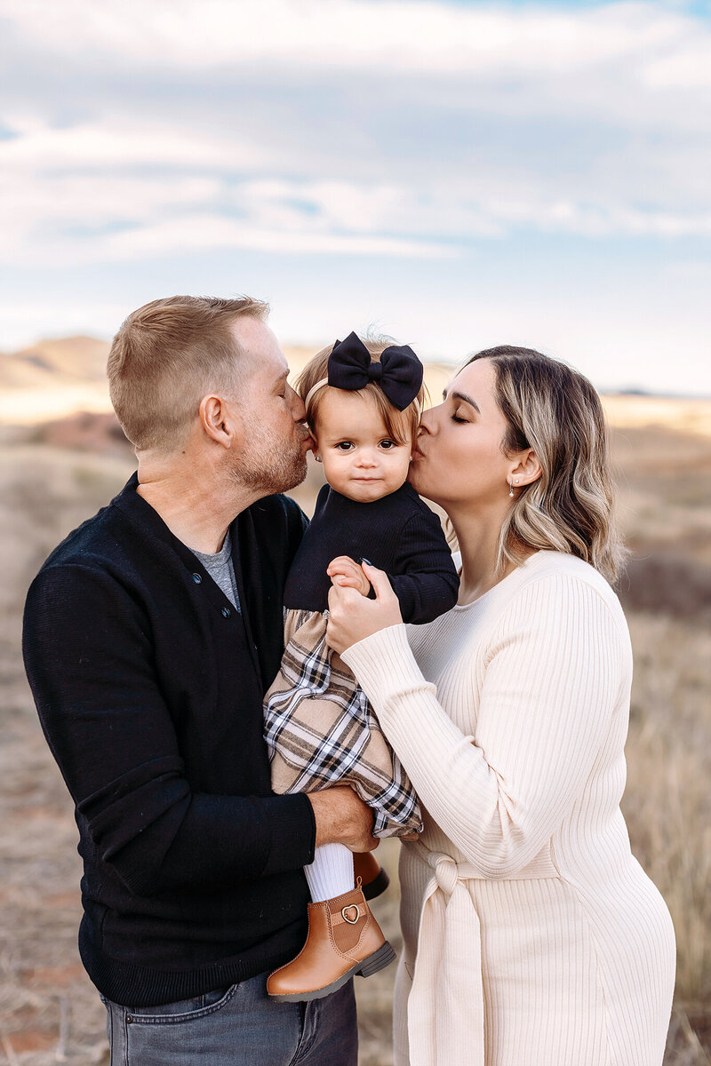 mom and dad holding their daughter and giving her kisses during their family photoshoot in erie colorado at sunset