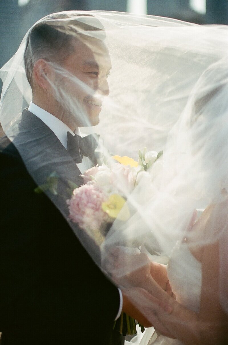 a film photograph of a bride and groom under a veil with sun coming through. candid.