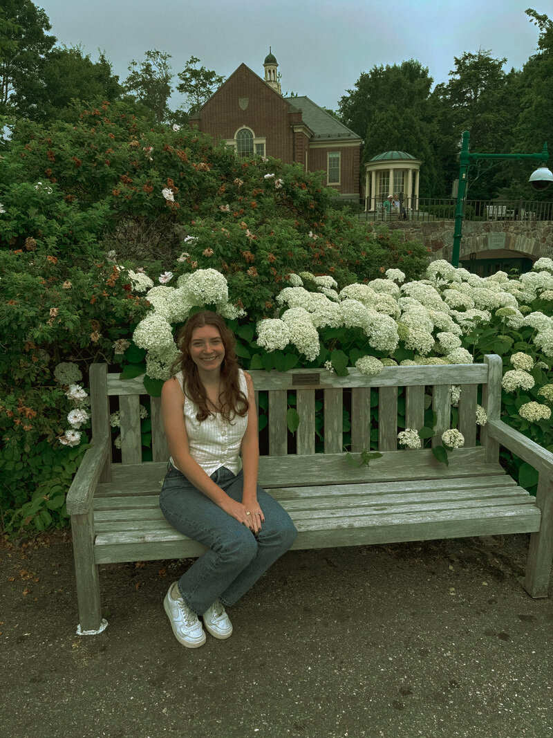 A woman can be seen smiling at the camera while sitting on a bench with flowers in the background