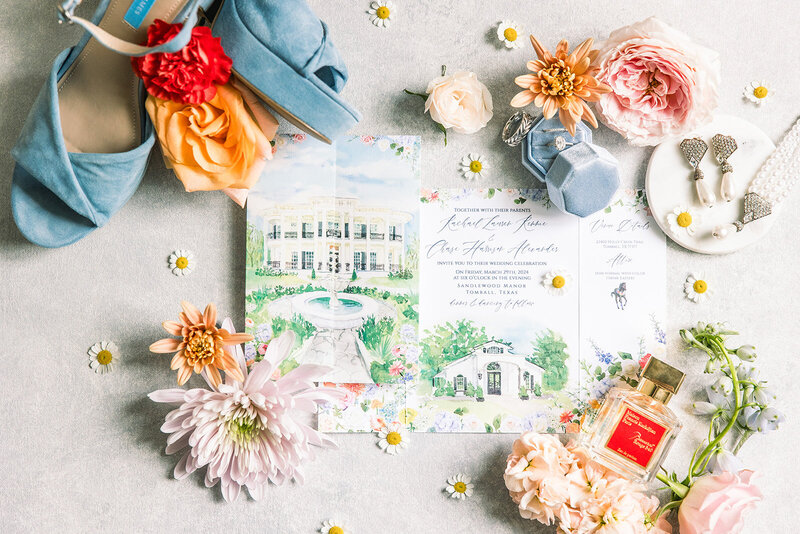 Flat lay of a watercolor wedding invitation suite surrounded by colorful flowers, blue shoes, jewelry, perfume, and a blue ring box