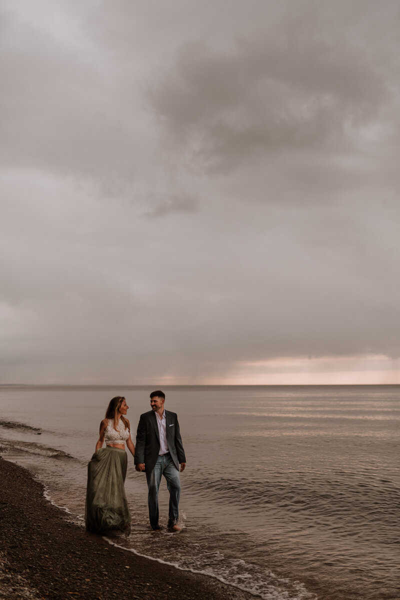 couple walking along shoreline holding hands at sunset