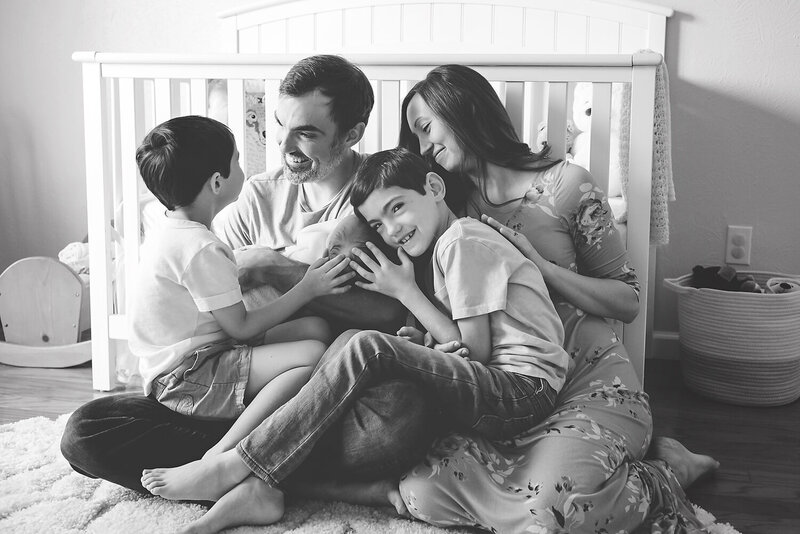 A family of five snuggling a newborn baby girl while sitting in front of the crib in her nursery.