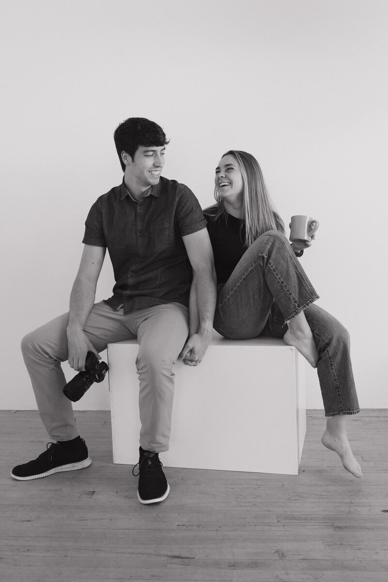 Savannah and Adam, a photographer and videographer at LeMay weddings based in Minneapolis Minnesota, sit on a studio block holding hands and smiling at each other. Adam holds his camera in one hand and Sav holds a cup of coffee in her hand. 