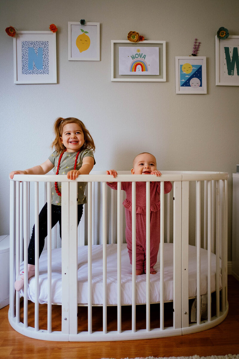Toddler girl and baby sister stand in a crib smiling.