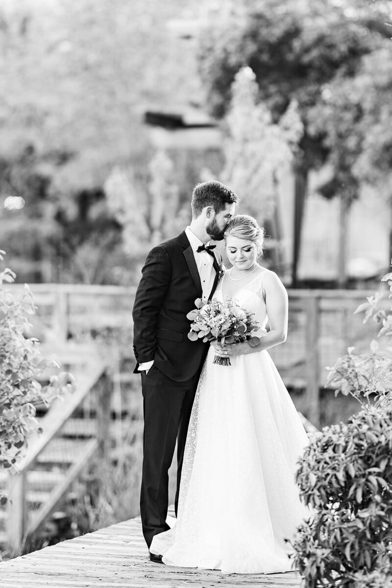 Elegant black & white wedding photograph of bride & groom with groom kissing the bride on top of her head while bride smiles down at her flowers. Photo taken by Chicago + Atlanta Photographer Ella Grace Richards of Forever Love Photography 